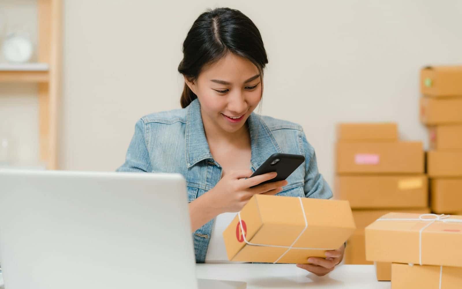 Dizüstü bilgisayar ve kargo kolilerinin yanında telefonundan siparişleri kontrol eden kadın
